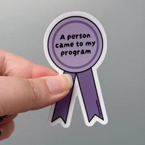 May include: A purple award ribbon sticker with a circular top and two downward-pointing tails. The center of the circle features the black text A person came to my program in a simple sans-serif font. The sticker has a thin white border.
