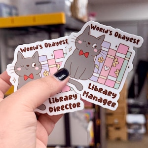 May include: Two stickers featuring a gray cat wearing a red bow tie, with the text "World's Okayest Library Manager" and "World's Okayest Library Director". The cat is drawn with a smiling face and is surrounded by colorful books and flowers.