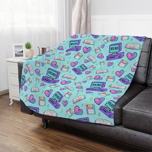 May include: A light blue throw blanket with a repeating pattern of retro computers with the word "READ" on the screen, hearts, open books, and floppy disks. The blanket is draped over a black couch.