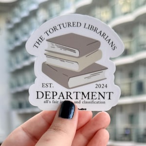 May include: A white sticker with the text "THE TORTURED LIBRARIANS" arched above a stack of three books. Below the books, the sticker reads "DEPARTMENT" and "EST. 2024". The text "all's fair in and classification" is also included.