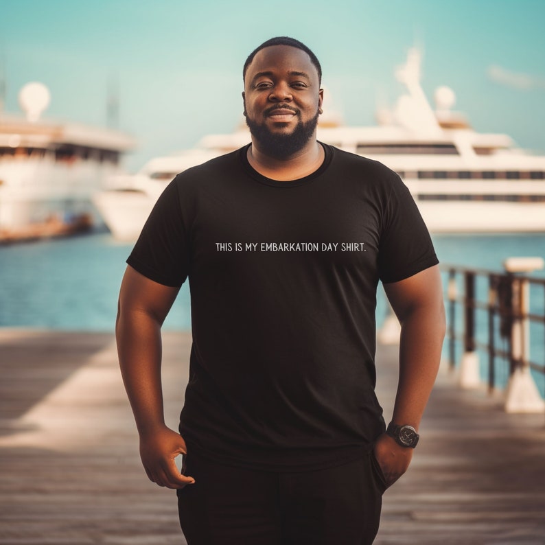 May include: A man wearing a black t-shirt with the text "THIS IS MY EMBARKATION DAY SHIRT."  He is standing on a wooden dock with a large white yacht in the background.