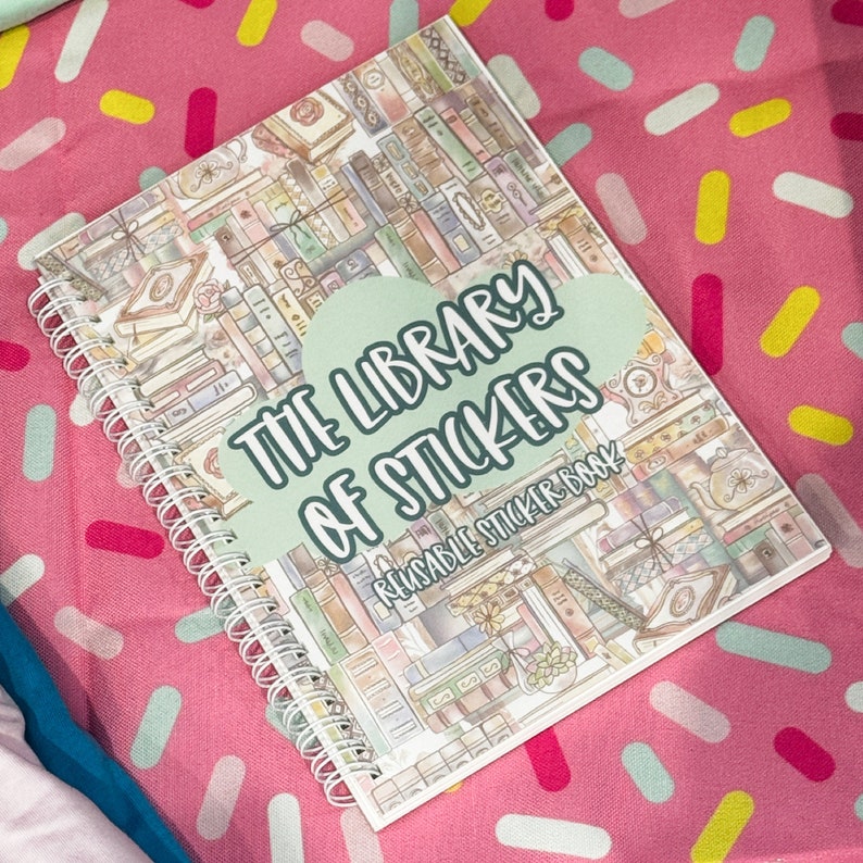 Pu&ograve; includere: Un libro di adesivi rilegato a spirale intitolato "The Library of Stickers". La copertina presenta un'illustrazione ad acquerello di una biblioteca con libri ed elementi decorativi. Il testo &egrave; in un carattere giocoso, su sfondo rosa.