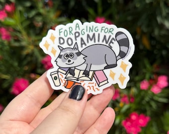 May include: A sticker featuring a cartoon raccoon lying on books with a coffee cup. The text on the sticker reads "Foraging for Dopamine" in a playful font, with yellow star accents.