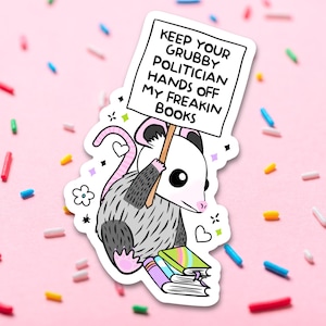 May include: A cartoon possum holding a sign that says "Keep your Grubby Politician Hands Off My Freakin Books" and sitting on a stack of books. The possum is gray and white with pink ears and nose.