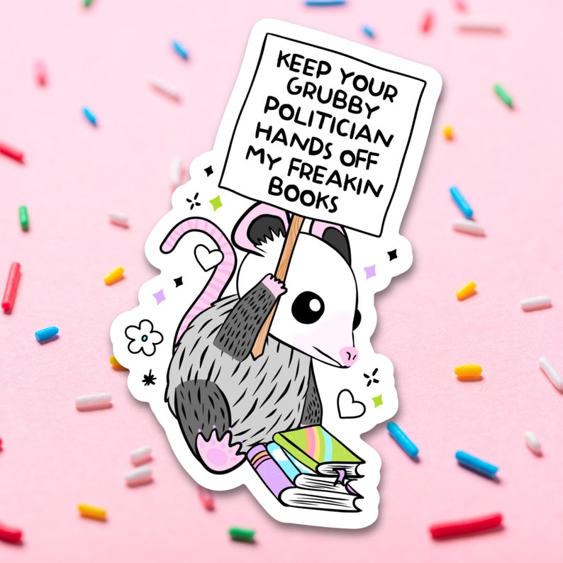 May include: A cartoon possum holding a sign that says "Keep your Grubby Politician Hands Off My Freakin Books" and sitting on a stack of books. The possum is gray and white with pink ears and nose.