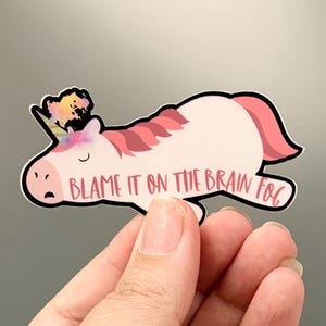 May include: A die-cut sticker featuring a light pink unicorn lying down with a closed eye and a rainbow-colored mane. The unicorn has a multi-colored horn and the text "BLAME IT ON THE BRAIN FOG" written in a dark pink script font across its body.