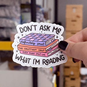 May include: A white sticker with the text "DON'T ASK ME WHAT I'M READING." The sticker features illustrations of books with the words "definitely smut," "THE SMUTTIEST SMUT," and "EVEN MORE SMUT LOL."