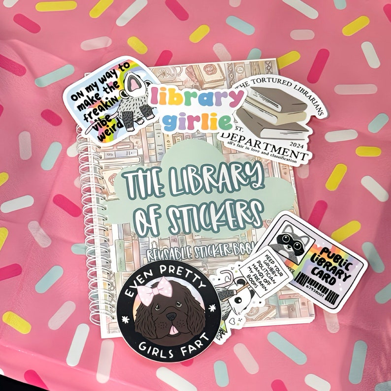Pu&ograve; includere: Un libro di adesivi a spirale intitolato "The Library of Stickers" con vari adesivi. Gli adesivi includono "Library Girlie", "The Tortured Librarians" e una tessera della biblioteca pubblica. Lo sfondo &egrave; una superficie rosa con coriandoli colorati.
