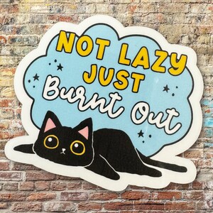 Może przedstawiać: Naklejka w kolorze niebieskim i białym z czarnym kotem i tekstem "Not Lazy Just Burnt Out" w ż&oacute;łtych literach.