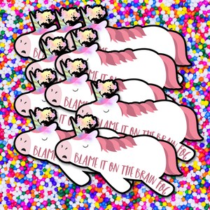 Op de afbeelding: Een verzameling witte eenhoornstickers met roze manen en staarten en een kleurrijke hoorn. De stickers hebben de tekst "BLAME IT ON THE BRAIN FOG" erop gedrukt. De stickers staan op een kleurrijke achtergrond.