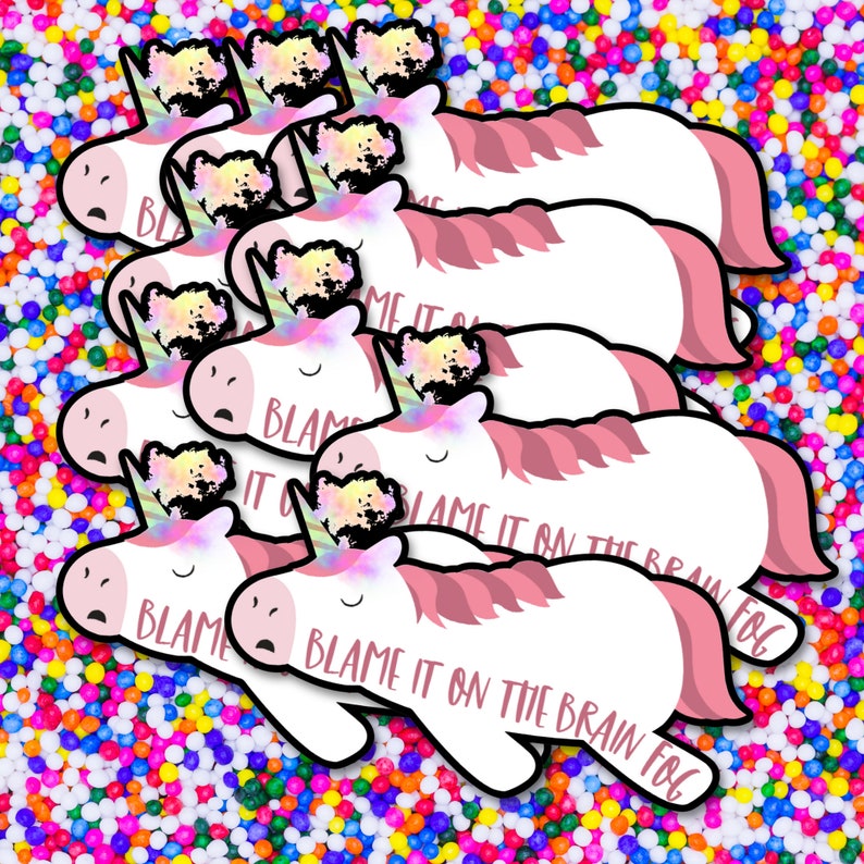 Op de afbeelding: Een verzameling witte eenhoornstickers met roze manen en staarten en een kleurrijke hoorn. De stickers hebben de tekst "BLAME IT ON THE BRAIN FOG" erop gedrukt. De stickers staan op een kleurrijke achtergrond.