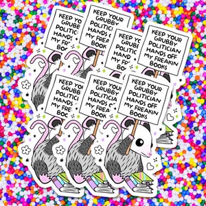 May include: A cartoon opossum holding a sign that reads "Keep your grubby politician hands off my freakin' books". The opossum is sitting on a stack of books. The background is a colorful confetti pattern.