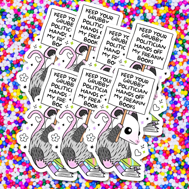 May include: A cartoon opossum holding a sign that reads "Keep your grubby politician hands off my freakin' books". The opossum is sitting on a stack of books. The background is a colorful confetti pattern.