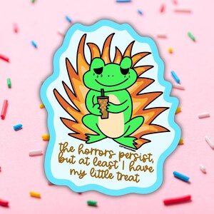 May include: A sticker featuring a green frog with a sad expression, holding a drink. The frog is set against a backdrop of orange flames and a light blue border. The text reads, "the horrors persist, but at least I have my little treat."
