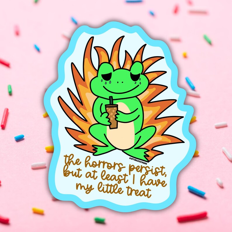 May include: A sticker featuring a green frog with a sad expression, holding a drink. The frog is set against a backdrop of orange flames and a light blue border. The text reads, "the horrors persist, but at least I have my little treat."