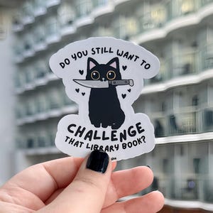 May include: A white sticker with a black cat illustration holding a knife. The text on the sticker reads "DO YOU STILL WANT TO CHALLENGE THAT LIBRARY BOOK?" in black, with heart accents.
