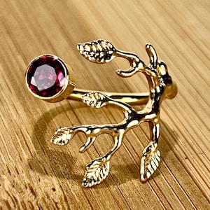May include: A gold-colored ring featuring a round, deep red gemstone and a branch-like design with leaf details. The ring is set against a wooden background, highlighting its intricate craftsmanship and natural inspiration.