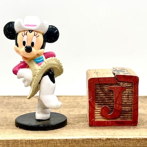 May include: A white and black cowgirl Minnie Mouse figurine wearing a pink and white cowboy hat and a tan vest. The figurine is standing on a black base. Next to the figurine is a wooden block with the letter J printed in red.