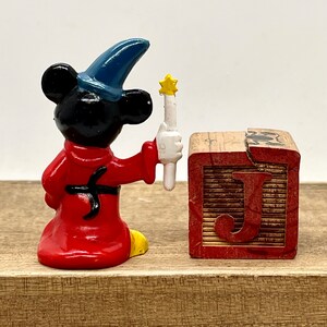 May include: A red and black Sorcerer Mickey Mouse figurine holding a white wand with a yellow star on top. The figurine is standing next to a wooden block with the letter 'J' painted on it in red.
