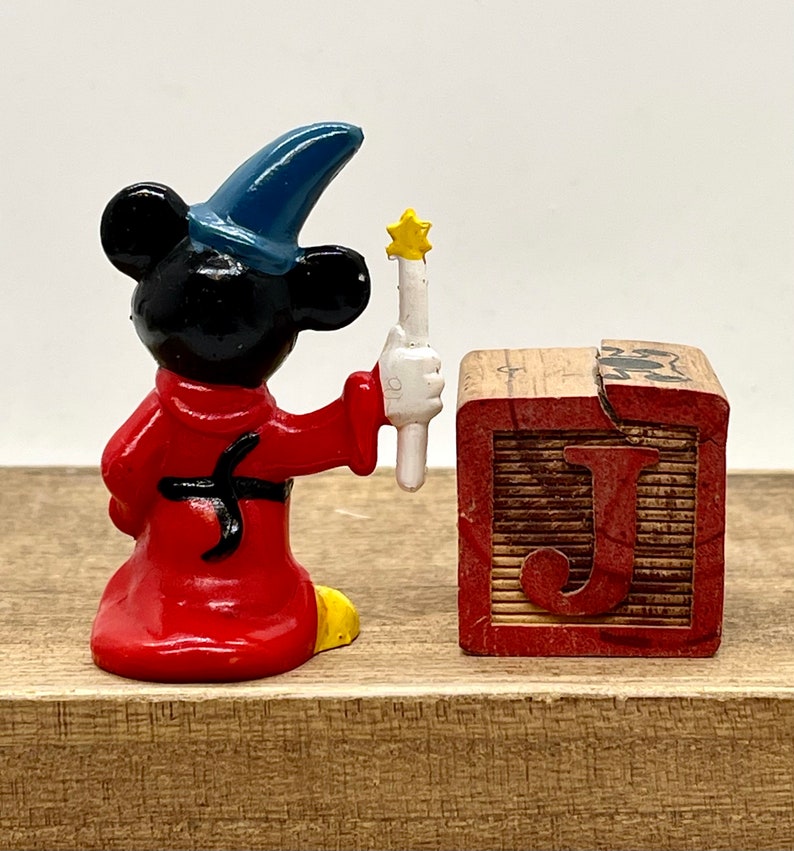 May include: A red and black Sorcerer Mickey Mouse figurine holding a white wand with a yellow star on top. The figurine is standing next to a wooden block with the letter 'J' painted on it in red.