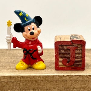 May include: A small plastic figurine of Mickey Mouse dressed as a sorcerer. He is wearing a blue hat with stars and a crescent moon, a red robe with a black sash, and yellow shoes. He is holding a white wand with a yellow star on top. The figurine is standing next to a wooden block with the letter "J" painted on it.