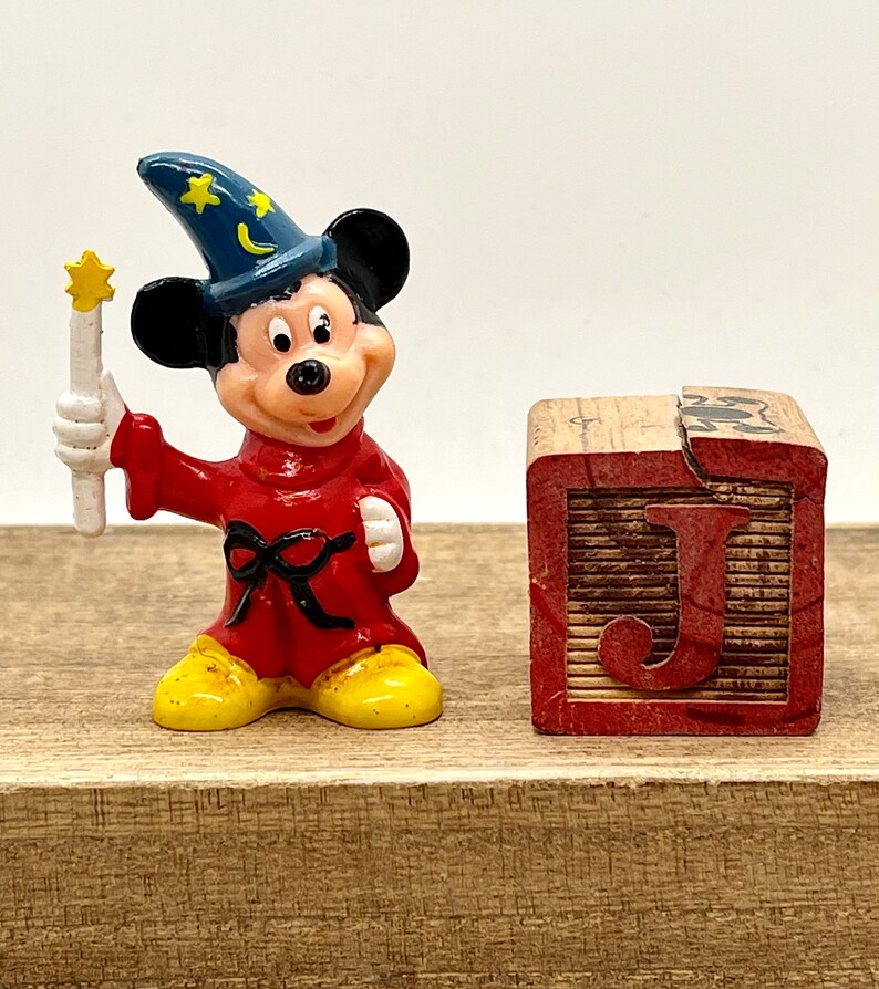 May include: A small plastic figurine of Mickey Mouse dressed as a sorcerer. He is wearing a blue hat with stars and a crescent moon, a red robe with a black sash, and yellow shoes. He is holding a white wand with a yellow star on top. The figurine is standing next to a wooden block with the letter "J" painted on it.