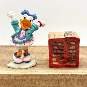 May include: A small, white duck figurine with blue hair and a pink polka dot dress, standing on a white base. The duck is holding its arms up in the air. A wooden block with the letter "J" painted on it is in the background.