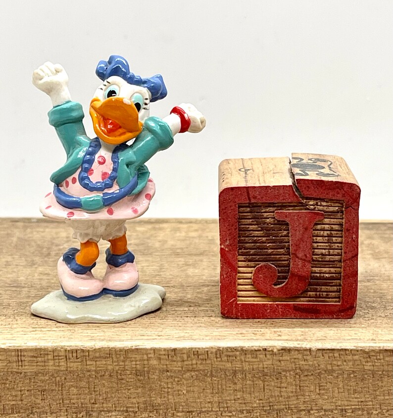 May include: A small, white duck figurine with blue hair and a pink polka dot dress, standing on a white base. The duck is holding its arms up in the air. A wooden block with the letter "J" painted on it is in the background.