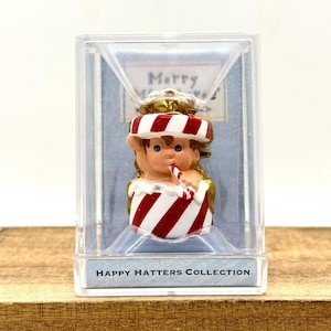 May include: A small figurine from the Happy Hatters Collection encased in a clear plastic box. The figurine depicts a character with brown hair wearing a red and white striped hat and sitting in a matching red and white striped container.
