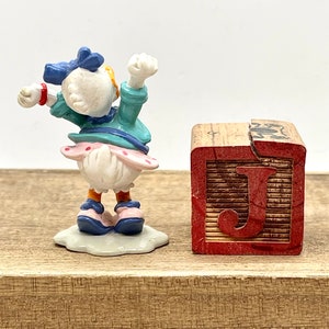 May include: A small figurine of a cartoon duck wearing a blue dress and a blue bow. The duck is standing on a white base and is holding its arms up in the air. The figurine is next to a wooden block with the letter "J" painted on it.