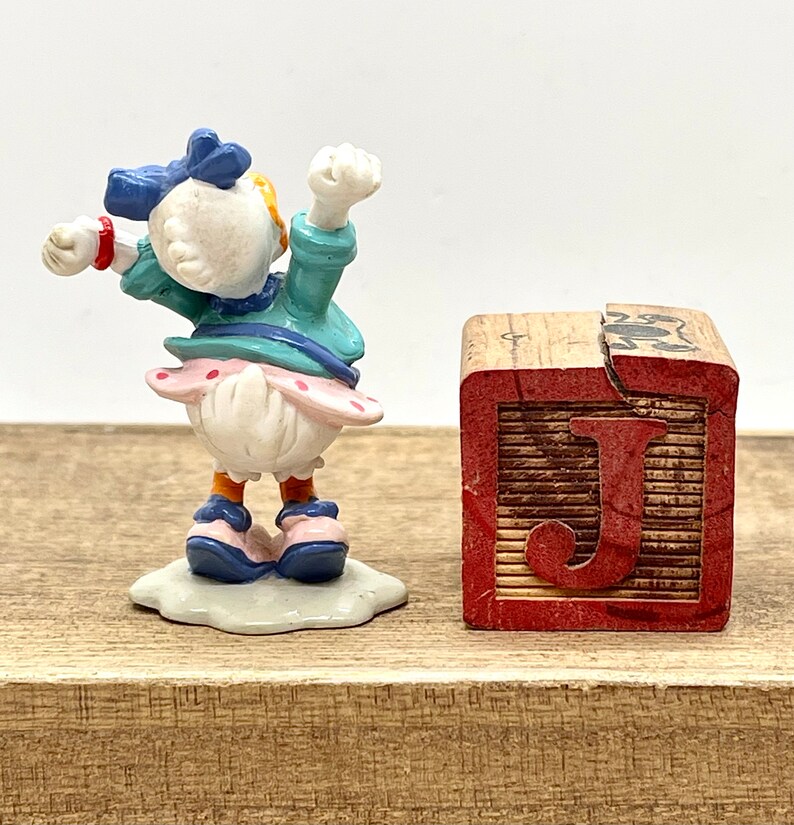 May include: A small figurine of a cartoon duck wearing a blue dress and a blue bow. The duck is standing on a white base and is holding its arms up in the air. The figurine is next to a wooden block with the letter "J" painted on it.