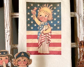 May include: A white tea towel with a vintage illustration of a child dressed as the Statue of Liberty holding a torch and scrolls. The child is standing in front of an American flag with red and white stripes and blue stars.