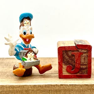 May include: A blue and white figurine of Donald Duck holding a lunchbox with two small mice inside. The figurine is standing next to a wooden block with the letter 'J' painted on it in red.