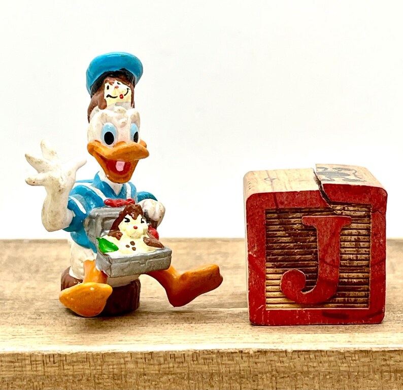 May include: A blue and white figurine of Donald Duck holding a lunchbox with two small mice inside. The figurine is standing next to a wooden block with the letter 'J' painted on it in red.