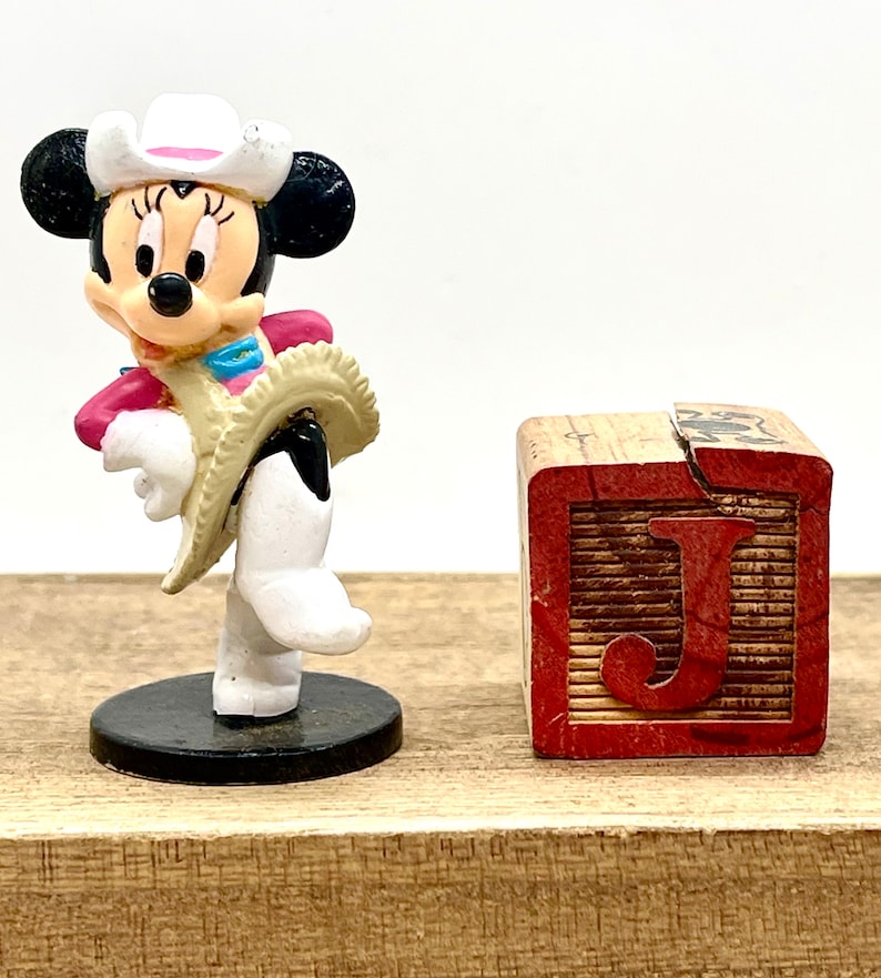 May include: A white and black cowgirl Minnie Mouse figurine wearing a pink and white cowboy hat and a tan vest. The figurine is standing on a black base. Next to the figurine is a wooden block with the letter J printed in red.