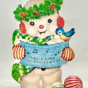 Puede incluir: Una tarjeta de Navidad vintage con un mu&ntilde;eco de nieve que lleva una corona verde y una bufanda azul, sosteniendo una hoja de m&uacute;sica con las palabras "Sing a song of Christmas". Un p&aacute;jaro azul est&aacute; posado en el brazo del mu&ntilde;eco de nieve, y hay dos adornos navide&ntilde;os rayados en primer plano.