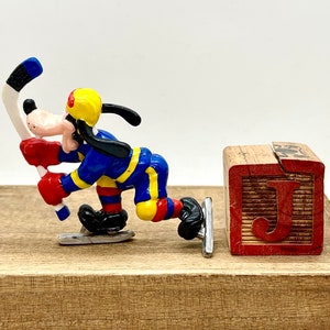 May include: A blue, yellow, and red Goofy figurine on a wooden surface holding a hockey stick. The figurine is wearing a hockey uniform and skates. A wooden block with the letter "J" is in the background.