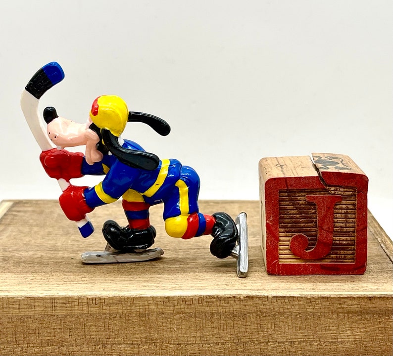 May include: A blue, yellow, and red Goofy figurine on a wooden surface holding a hockey stick. The figurine is wearing a hockey uniform and skates. A wooden block with the letter "J" is in the background.