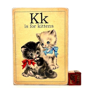 May include: Vintage educational card featuring two kittens with blue and red bows, with the text "Kk is for kittens." A small wooden block with the letter "J" is visible in the corner. The card has a cream-colored background.
