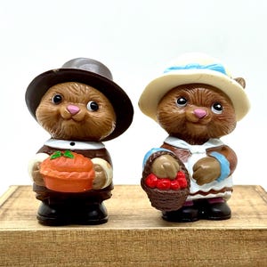 May include: Two small brown animal figurines. The left figure wears a dark brown hat and holds an orange pie. The right figure wears a cream-colored hat with a blue ribbon and holds a brown basket filled with small red berries.