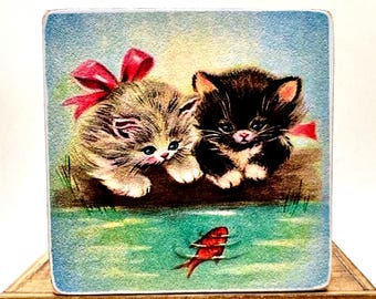 May include: Two kittens, one gray and white with a red bow, and one black and white, are looking at a red goldfish swimming in a blue pond.