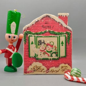 Puede incluir: Una casa de Navidad roja y blanca con una corona verde y una ventana que muestra dos mu&ntilde;ecos de nieve. La casa tiene un techo blanco con efecto de nieve y el texto "Hi There!" en la parte delantera.