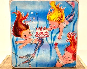 May include: A wooden block featuring a vintage illustration of three mermaids celebrating a birthday. The mermaids have flowing red and yellow hair, and are swimming in blue water. One mermaid holds a cake.