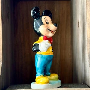 May include: A vintage Mickey Mouse figurine. The figure is wearing a yellow jacket, blue pants, and yellow shoes. It has a red bow tie and white gloves. The figurine is standing on a small, light blue base.