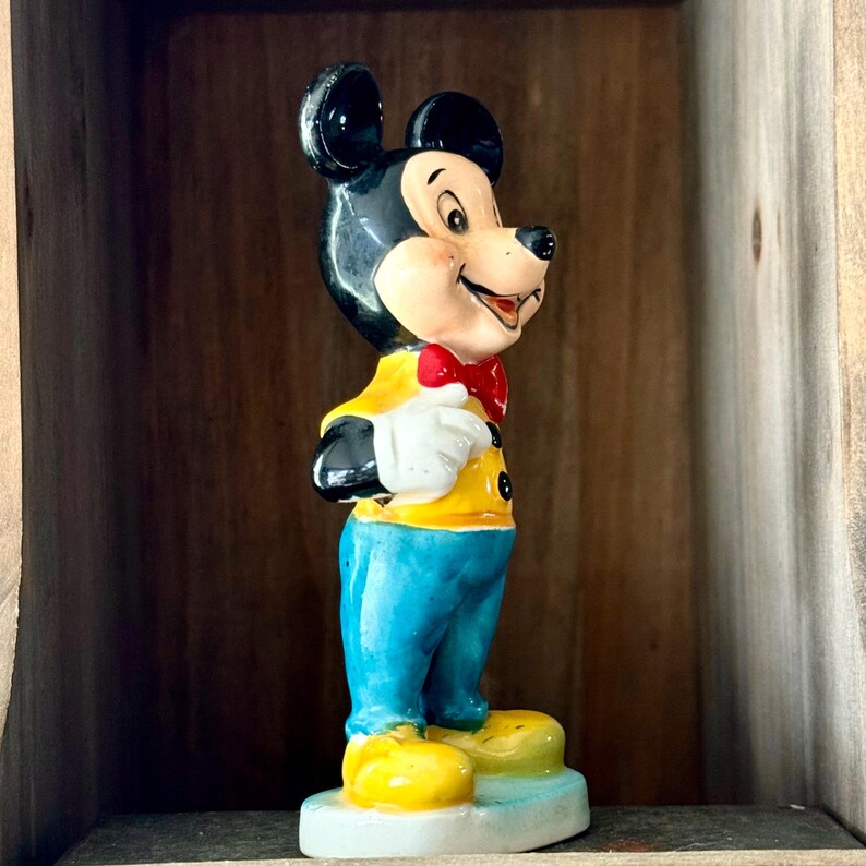 May include: A vintage Mickey Mouse figurine. The figure is wearing a yellow jacket, blue pants, and yellow shoes. It has a red bow tie and white gloves. The figurine is standing on a small, light blue base.