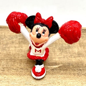 Może przedstawiać: Figurkę cheerleadera Minnie Mouse w czerwono-białych kolorach trzymająca czerwone pompony. Ma na sobie czerwoną sp&oacute;dniczkę z białym "M".