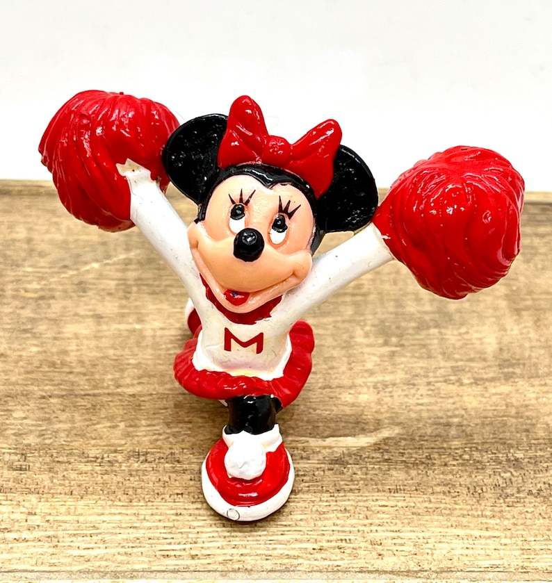 Może przedstawiać: Figurkę cheerleadera Minnie Mouse w czerwono-białych kolorach trzymająca czerwone pompony. Ma na sobie czerwoną sp&oacute;dniczkę z białym "M".