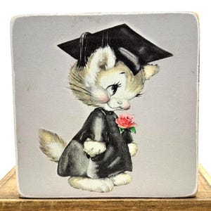 May include: A square, light gray block featuring a vintage illustration of a cat in a graduation cap and gown. The cat is light brown and white with pink cheeks, holding a red rose. The cat is wearing a black graduation cap and gown.