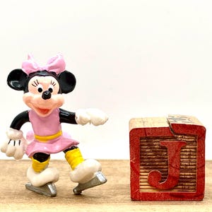 May include: A pink and white figurine of Minnie Mouse ice skating. She is wearing a pink dress with a yellow belt and a pink bow. She is standing on a wooden block with the letter "J" on it.