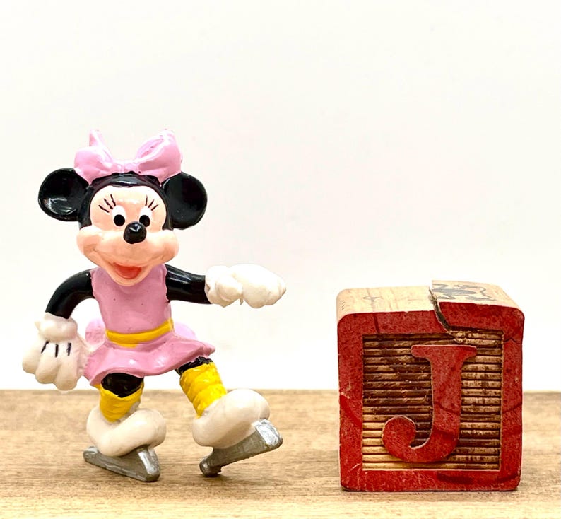 May include: A pink and white figurine of Minnie Mouse ice skating. She is wearing a pink dress with a yellow belt and a pink bow. She is standing on a wooden block with the letter "J" on it.